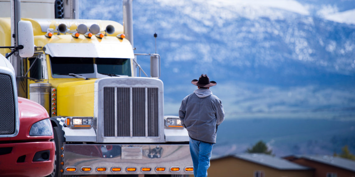 The truck driver goes to his bright yellow attractive impressive customized big rig semi truck parked in the parking lot on a picturesque backdrop of snow-capped mountain ranges.