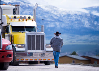 The truck driver goes to his bright yellow attractive impressive customized big rig semi truck parked in the parking lot on a picturesque backdrop of snow-capped mountain ranges.