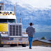 The truck driver goes to his bright yellow attractive impressive customized big rig semi truck parked in the parking lot on a picturesque backdrop of snow-capped mountain ranges.
