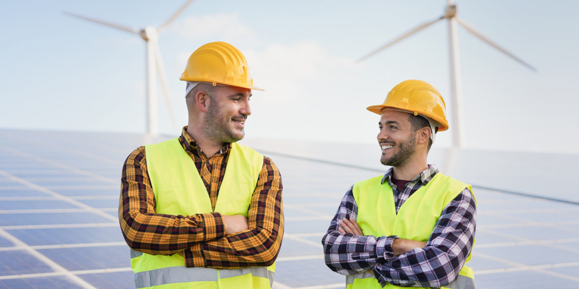 Worker men at solar power station - Solar panels with wind turbines in background - Green energy renewable concept