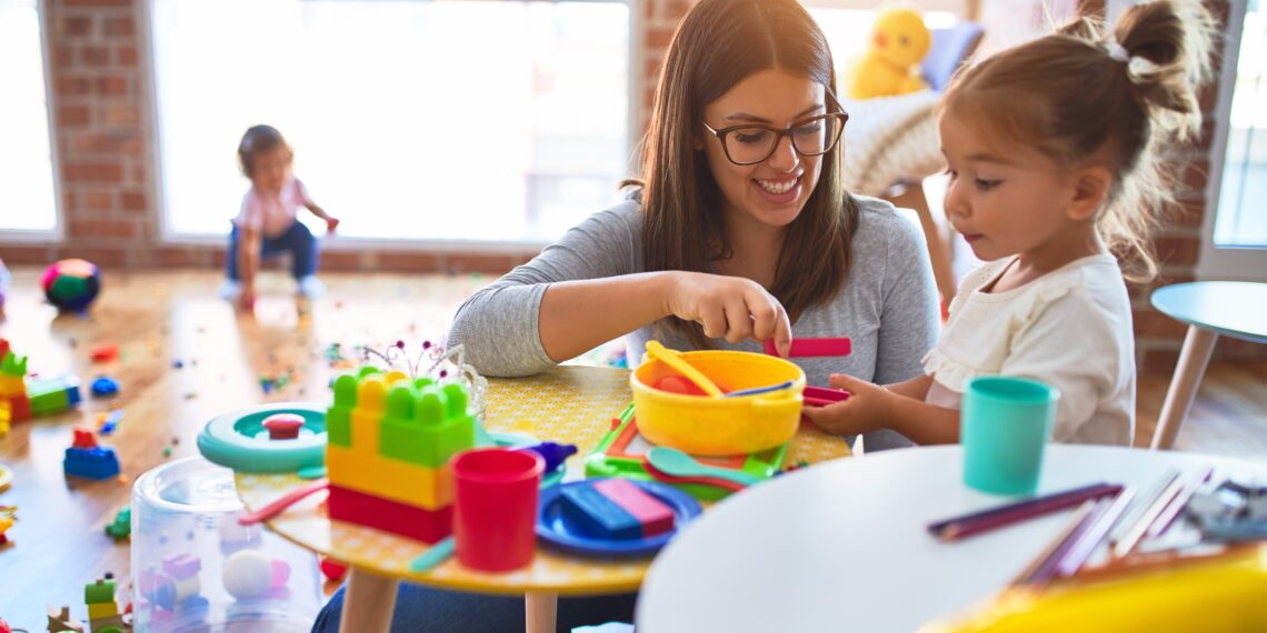 Young beautiful teacher and toddlers playing on the table with lots of toys at kindergarten