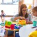 Young beautiful teacher and toddlers playing on the table with lots of toys at kindergarten