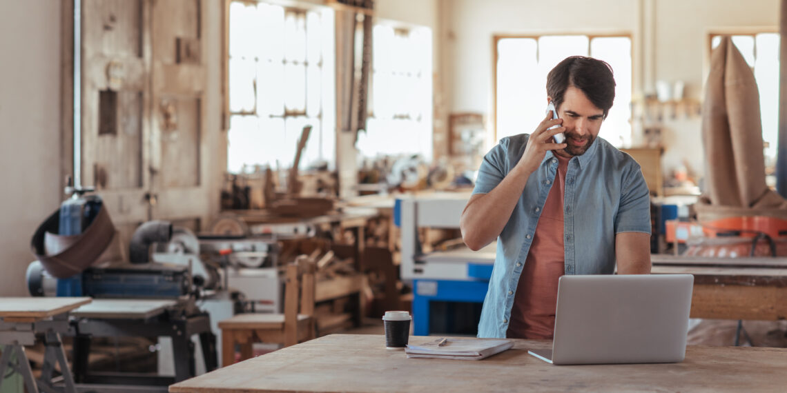 Skilled young craftsman standing at a workbench in his large workshop working online with a laptop and talking on a cellphone