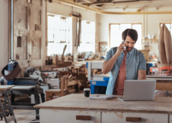 Skilled young craftsman standing at a workbench in his large workshop working online with a laptop and talking on a cellphone
