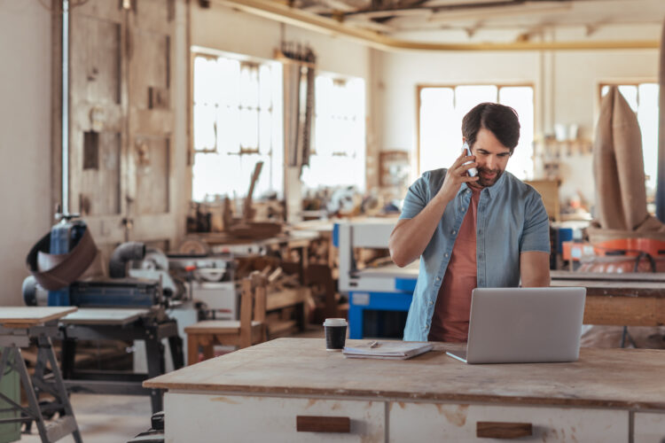 Skilled young craftsman standing at a workbench in his large workshop working online with a laptop and talking on a cellphone