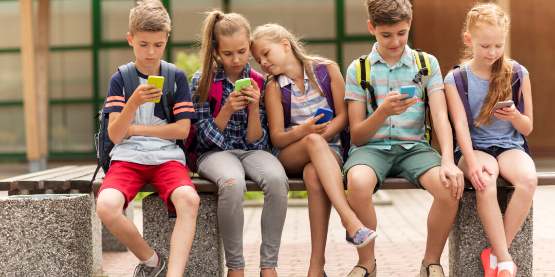 primary education, friendship, childhood, technology and people concept - group of happy elementary school students with smartphones and backpacks sitting on bench outdoors