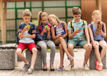 primary education, friendship, childhood, technology and people concept - group of happy elementary school students with smartphones and backpacks sitting on bench outdoors