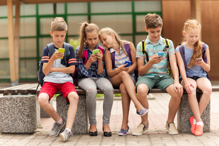 primary education, friendship, childhood, technology and people concept - group of happy elementary school students with smartphones and backpacks sitting on bench outdoors