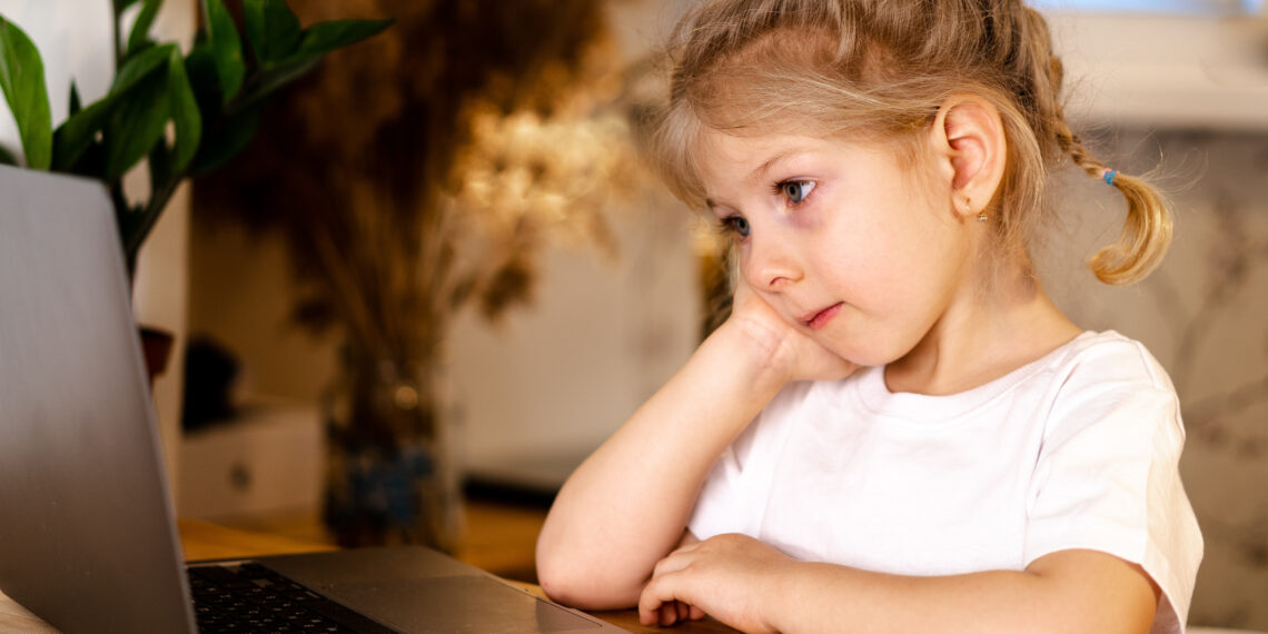 little pensive blonde girl sitting in front of a laptop with a sad face, the concept of home schooling and loneliness
