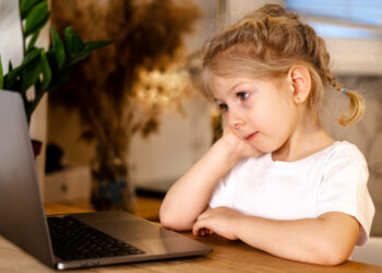 little pensive blonde girl sitting in front of a laptop with a sad face, the concept of home schooling and loneliness