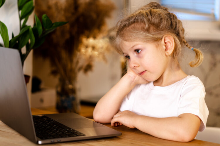 little pensive blonde girl sitting in front of a laptop with a sad face, the concept of home schooling and loneliness