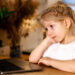 little pensive blonde girl sitting in front of a laptop with a sad face, the concept of home schooling and loneliness