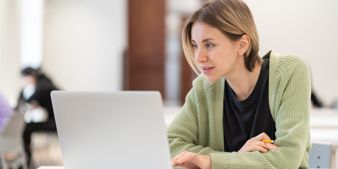 Smiling middle-aged woman studying online on laptop computer while sitting in classroom, doing second degree as mature student. Happy 45s female attending online professional development course