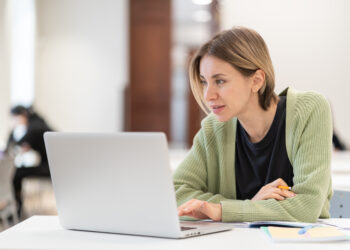 Smiling middle-aged woman studying online on laptop computer while sitting in classroom, doing second degree as mature student. Happy 45s female attending online professional development course