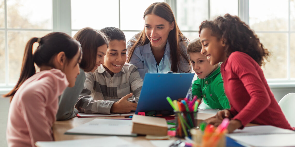 Happy Diverse School Children And Teacher Woman Having Class Sitting At Desk In Classroom At School. Modern Education And Knowledge Concept. Selective Focus