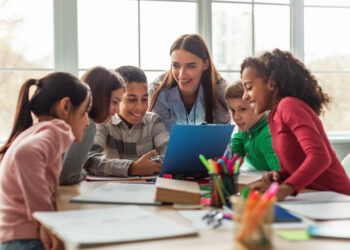 Happy Diverse School Children And Teacher Woman Having Class Sitting At Desk In Classroom At School. Modern Education And Knowledge Concept. Selective Focus