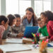 Happy Diverse School Children And Teacher Woman Having Class Sitting At Desk In Classroom At School. Modern Education And Knowledge Concept. Selective Focus